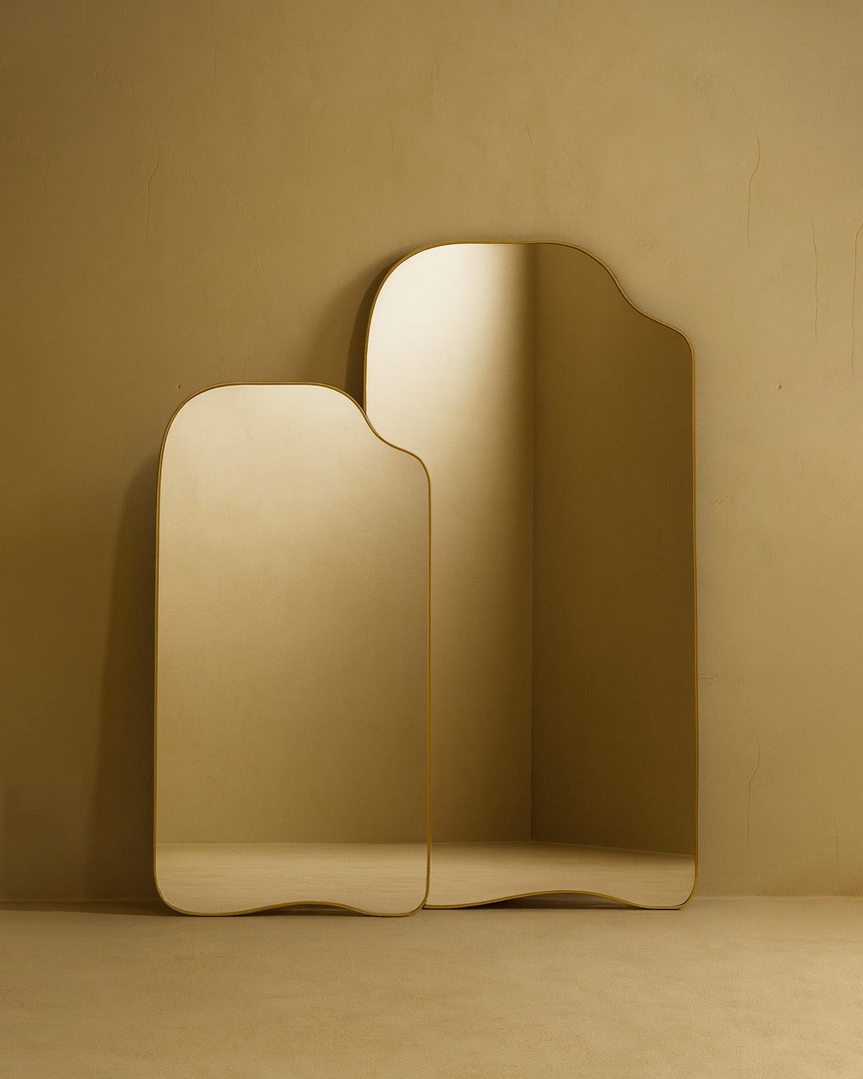 Asymmetrical full-length mirrors in two sizes with organic shapes and thin brass-colored frames, shown standing against a warm neutral wall on the floor.