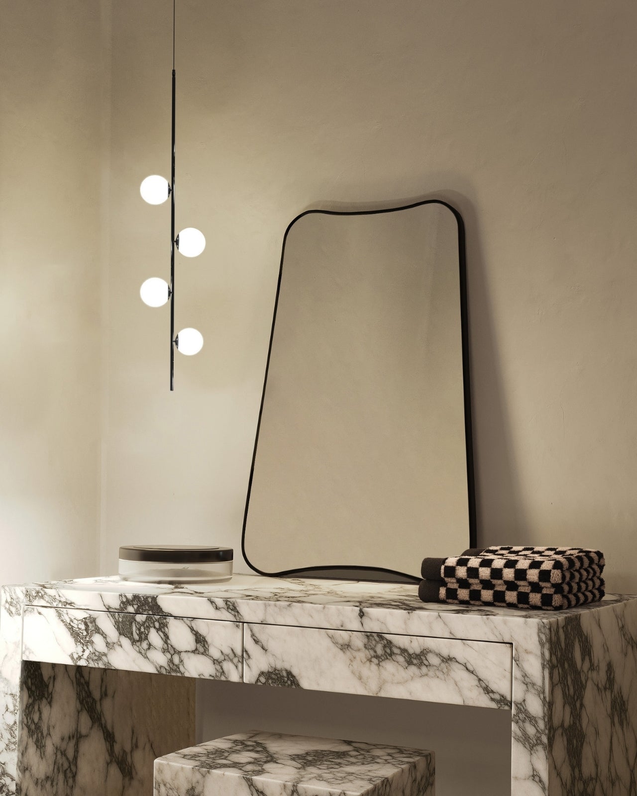 Modern vanity with a black irregular mirror, marble console table, stacked towels, and a vertical globe wall light.
