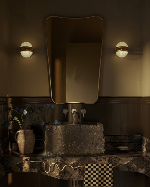 Butterfly-shaped wall mirror with curved edges and a thin brass frame mounted above a stone bathroom sink, flanked by two wall sconces.