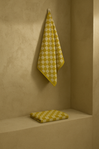 Yellow mustard and white checkered towel hanging on a wall hook, with a folded matching towel placed on a shelf below, highlighting the geometric pattern and terry texture.