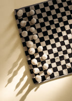 Black and white checkered towel styled with white sculptural chess pieces, photographed from above on a neutral background.