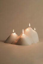 White sculptural candle with an organic, mountain-inspired shape and multiple lit cotton wicks, placed on a flat surface against a neutral background.