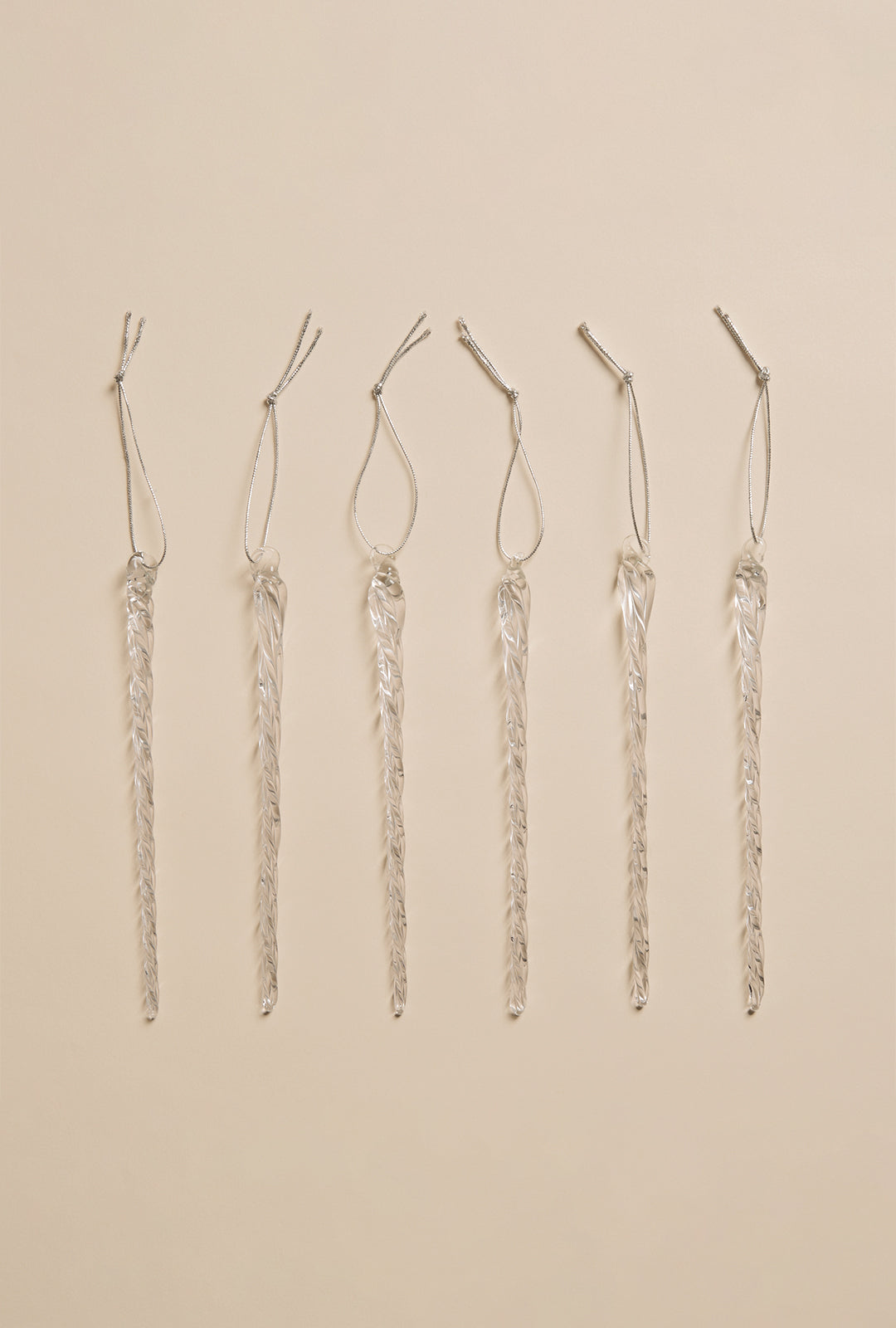 Six clear glass icicle ornaments with hanging loops arranged in a straight row on a neutral background.