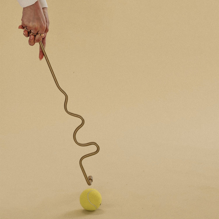 Hand holding a wavy brass towel bar touching a tennis ball on a neutral background.