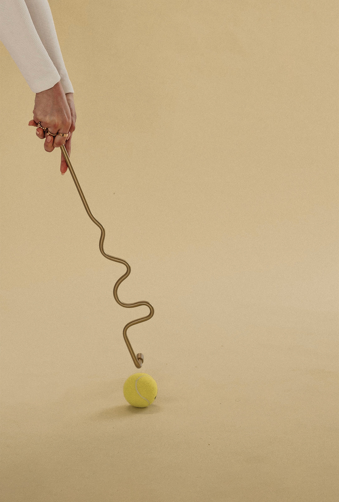 Hand holding a wavy brass towel bar touching a tennis ball on a neutral background.