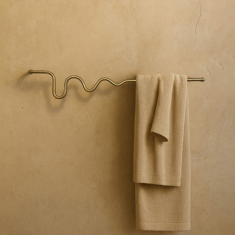 Wall-mounted towel bar with a wavy brass-colored design, holding a folded beige towel against a textured neutral wall.