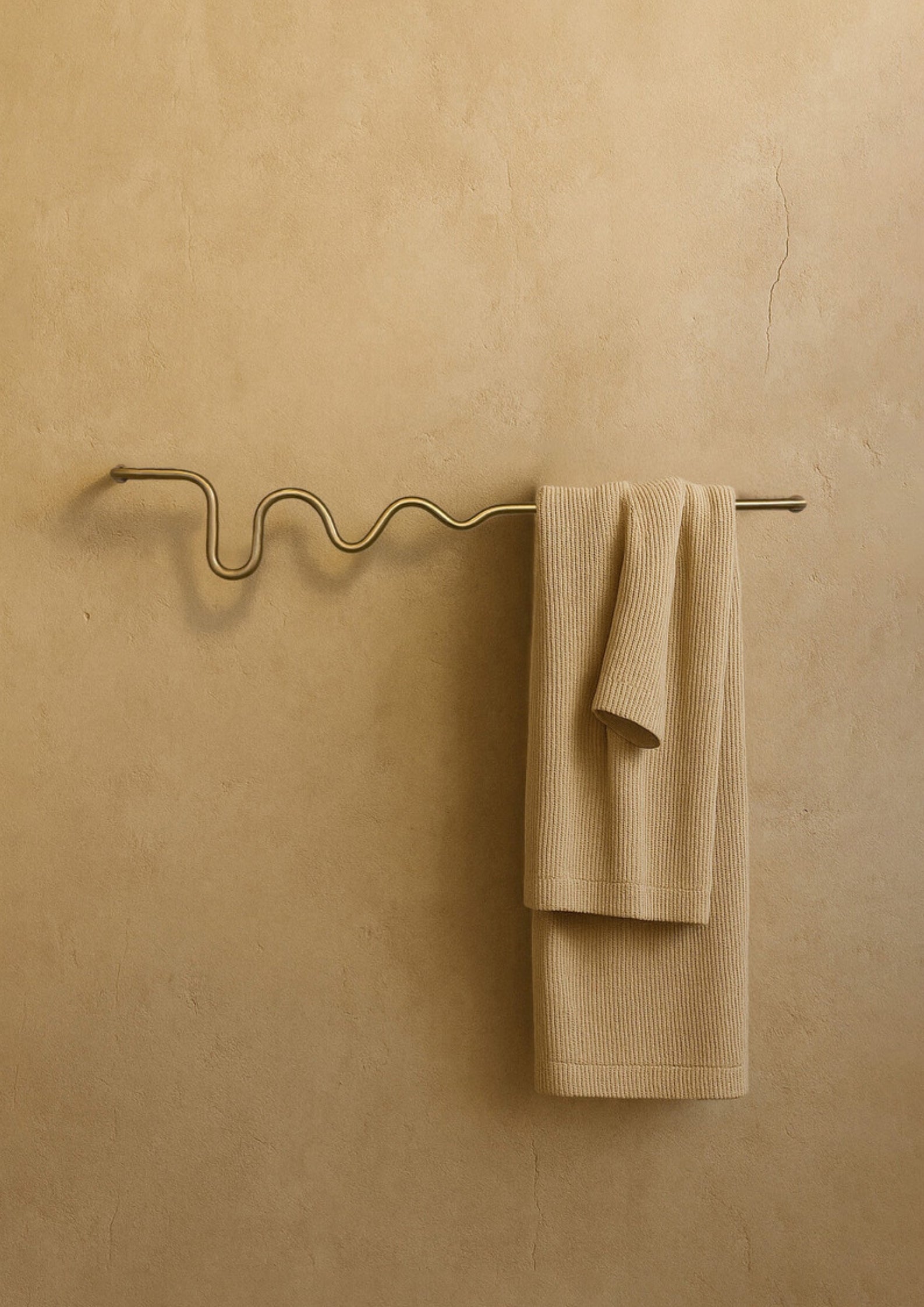 Wall-mounted towel bar with a wavy brass-colored design, holding a folded beige towel against a textured neutral wall.