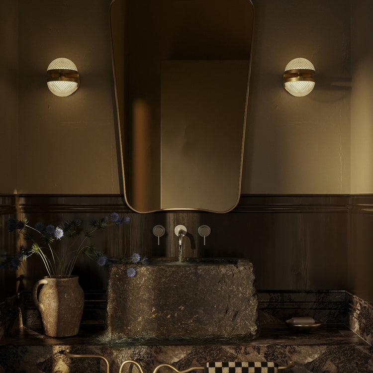 Moody bathroom with an irregular wall mirror, globe sconces, a stone vessel sink, and a brass wavy towel bar with a checkered towel.
