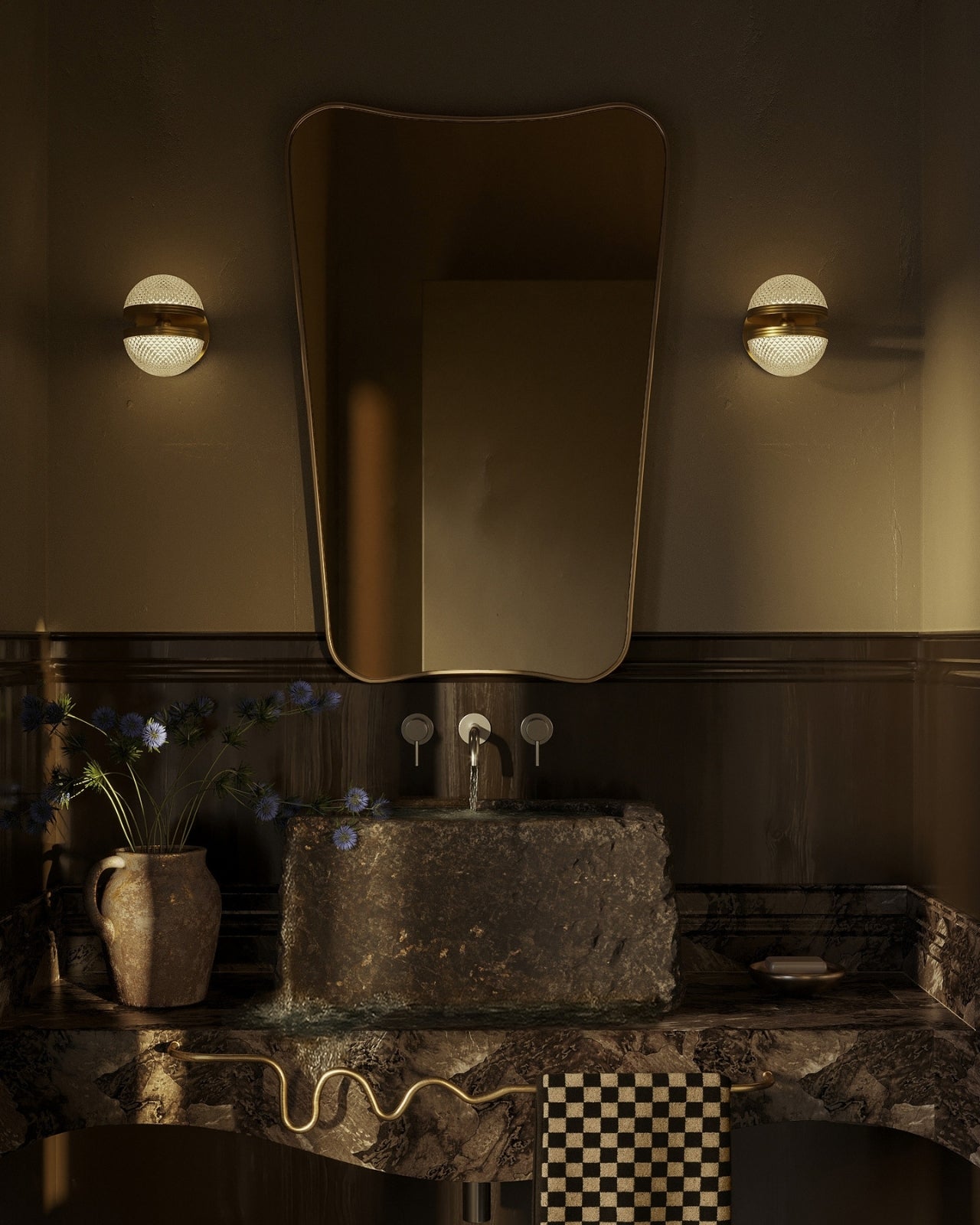 Moody bathroom with an irregular wall mirror, globe sconces, a stone vessel sink, and a brass wavy towel bar with a checkered towel.