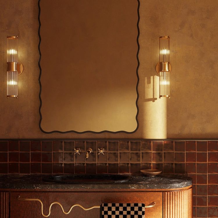 Wavy-edged mirror above a bathroom sink with wall lights and a towel hanging from a curved brass towel bar.