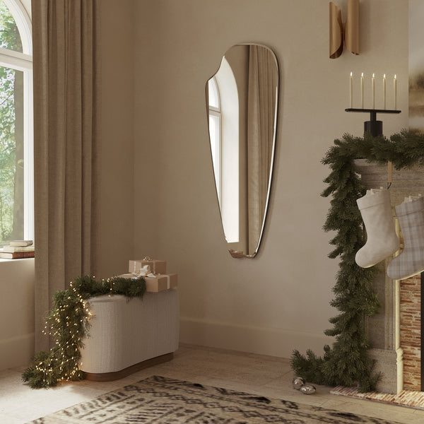 Frameless elongated mirror with an organic, sculptural silhouette styled in a warm living space with holiday greenery, candles, and soft natural light.