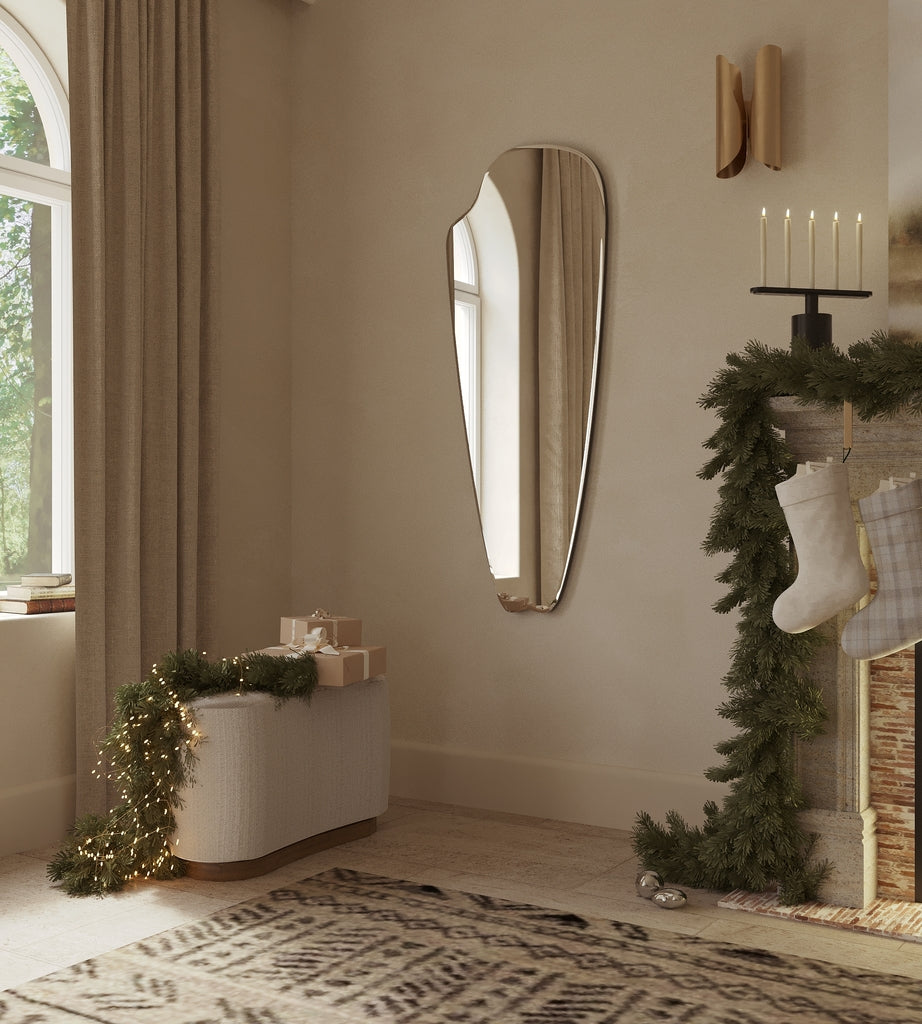 Frameless elongated mirror with an organic, sculptural silhouette styled in a warm living space with holiday greenery, candles, and soft natural light.