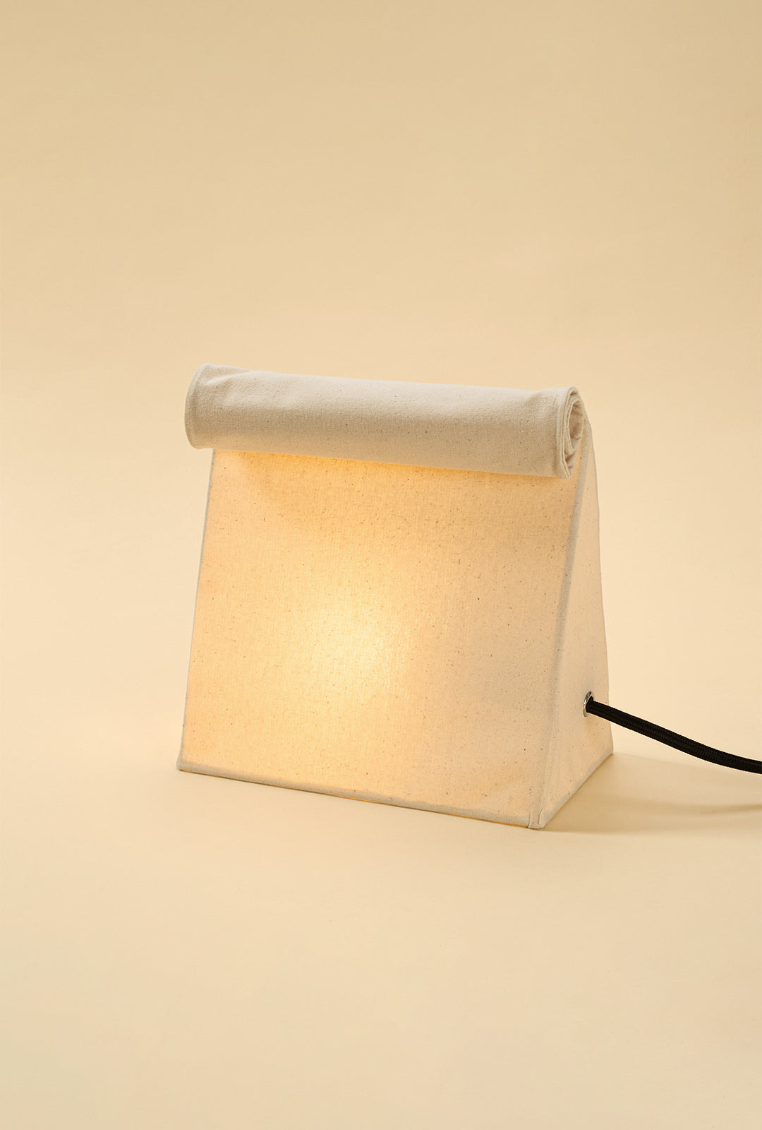 Illuminated linen lunch bag–style table lamp with a rolled top, emitting a warm ambient glow against a soft neutral background.