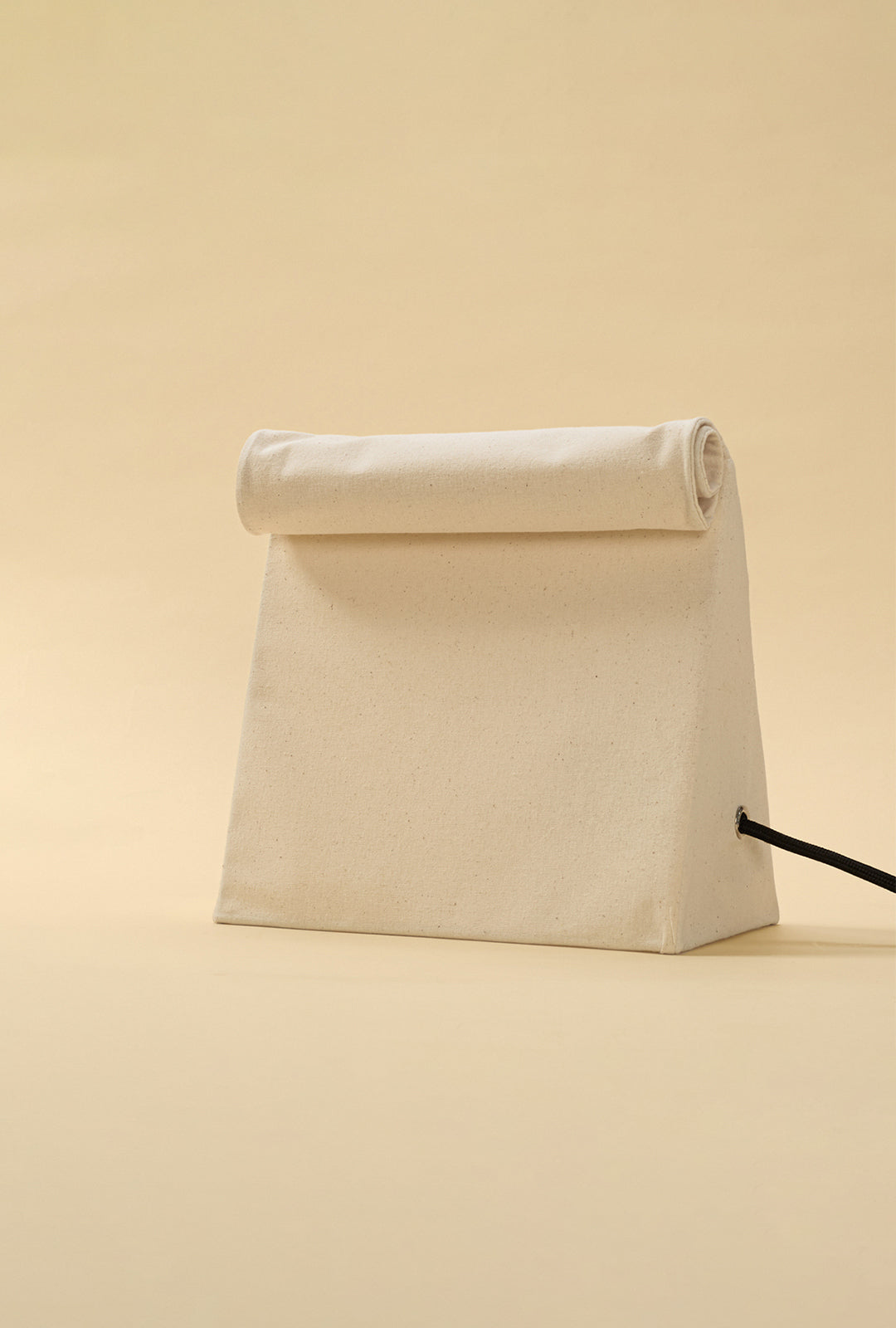 Minimal linen lunch bag–style table lamp with a rolled top and visible black power cord, set against a warm neutral background.