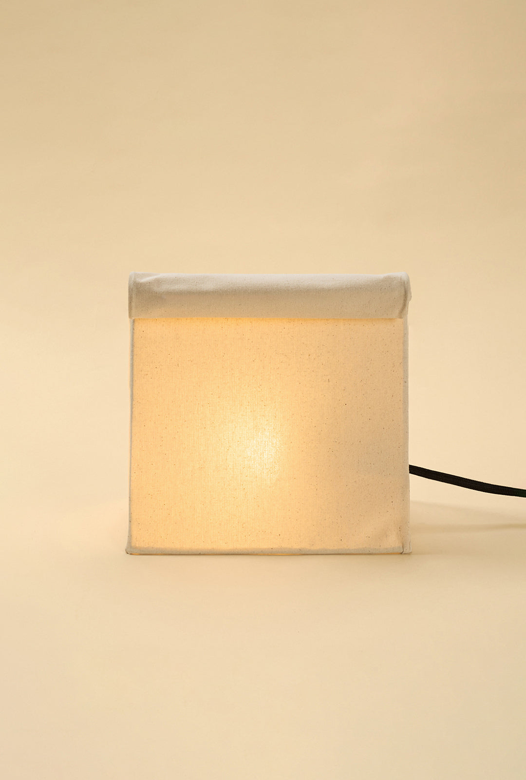 Lit linen table lamp with a lunch bag–inspired silhouette and rolled top, softly glowing against a warm neutral background.