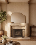 Asymmetrical brass-framed mirror mounted above a stone fireplace in a neutral interior setting.
