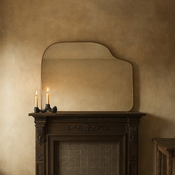 Asymmetrical brass-framed mirror mounted above a fireplace mantel, with two lit candles on the mantel below.