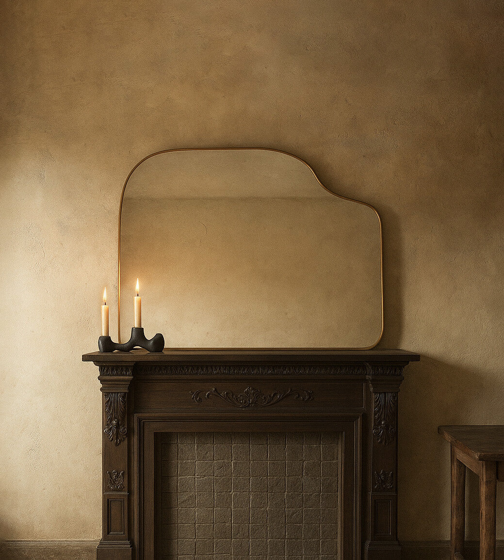 Asymmetrical brass-framed mirror mounted above a fireplace mantel, with two lit candles on the mantel below.