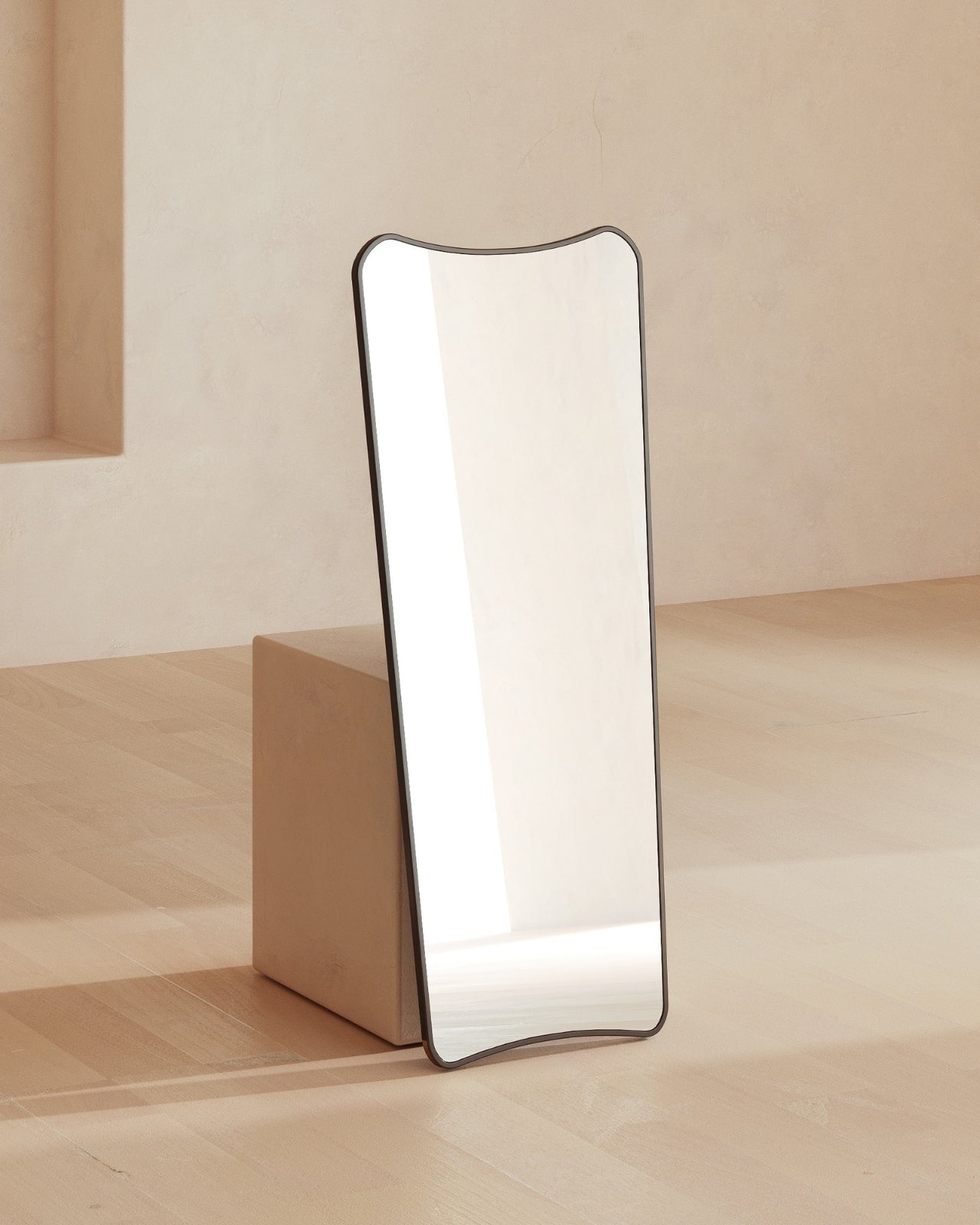 Black framed full-length mirror with a softly curved, asymmetrical silhouette, leaning against a low block in a minimal, light-toned interior.