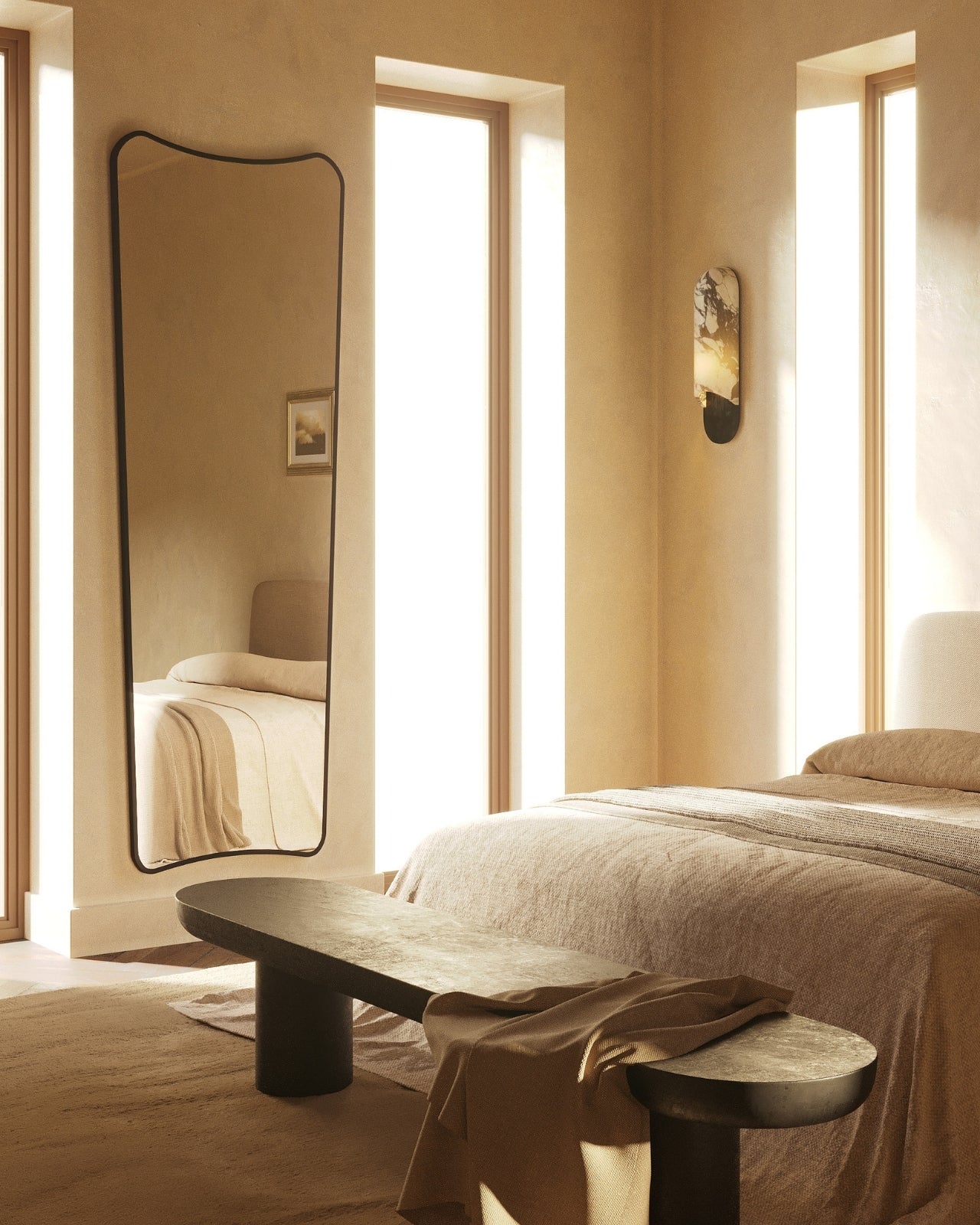 Minimalist bedroom with a low upholstered bed, textured area rug, dark wood bench, and a tall wavy-edge mirror reflecting soft natural light from narrow windows.