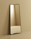 Tall full-length mirror with softly curved edges leaning against a wall, reflecting a minimal interior corner.
