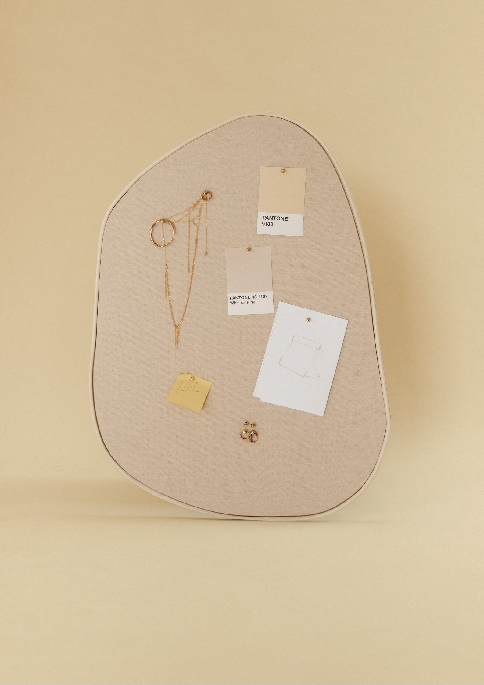 Asymmetrical fabric-covered pinboard with a beige outer frame, featuring pinned Pantone color cards, a small sketch, and gold jewelry.