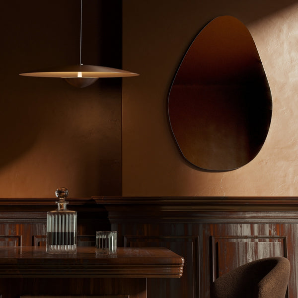 Pebble-shaped frameless wall mirror mounted above a wooden desk in a dimly lit interior.