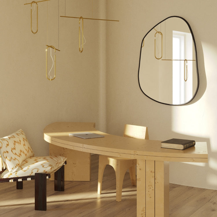 Asymmetrical wall mirror mounted above a sculptural wooden desk in a minimalist workspace with hanging brass light fixtures and natural wood flooring.
