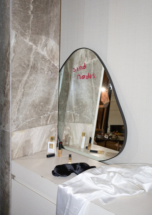 Vanity table with an asymmetrical mirror, cosmetics, and fabric draped on top.