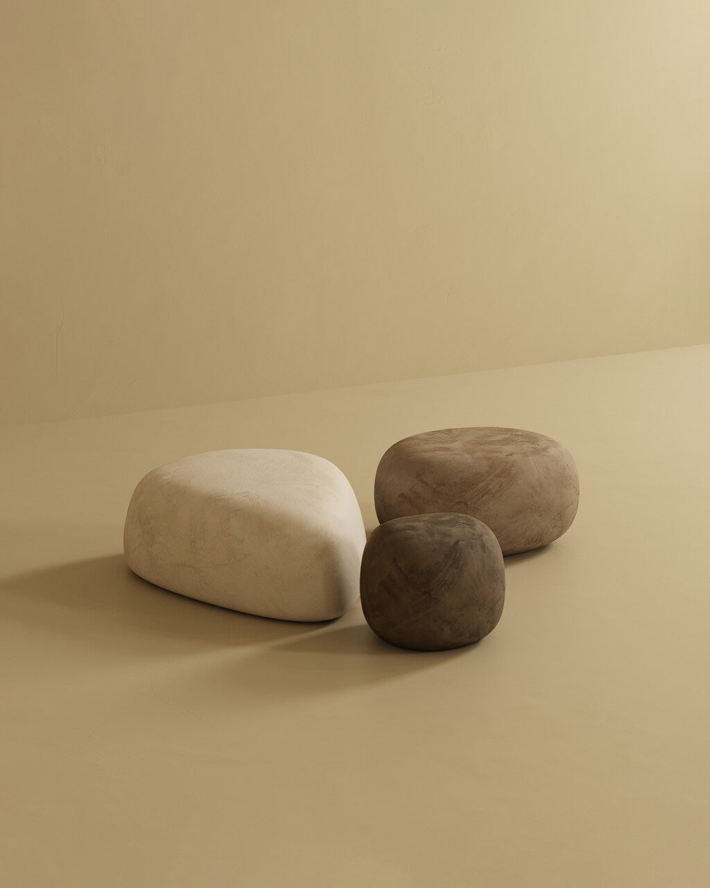 Three pebble-shaped ottomans in soft neutral colors, including one large off-white ottoman and two smaller brown ottomans, arranged together on a minimalist beige studio background.