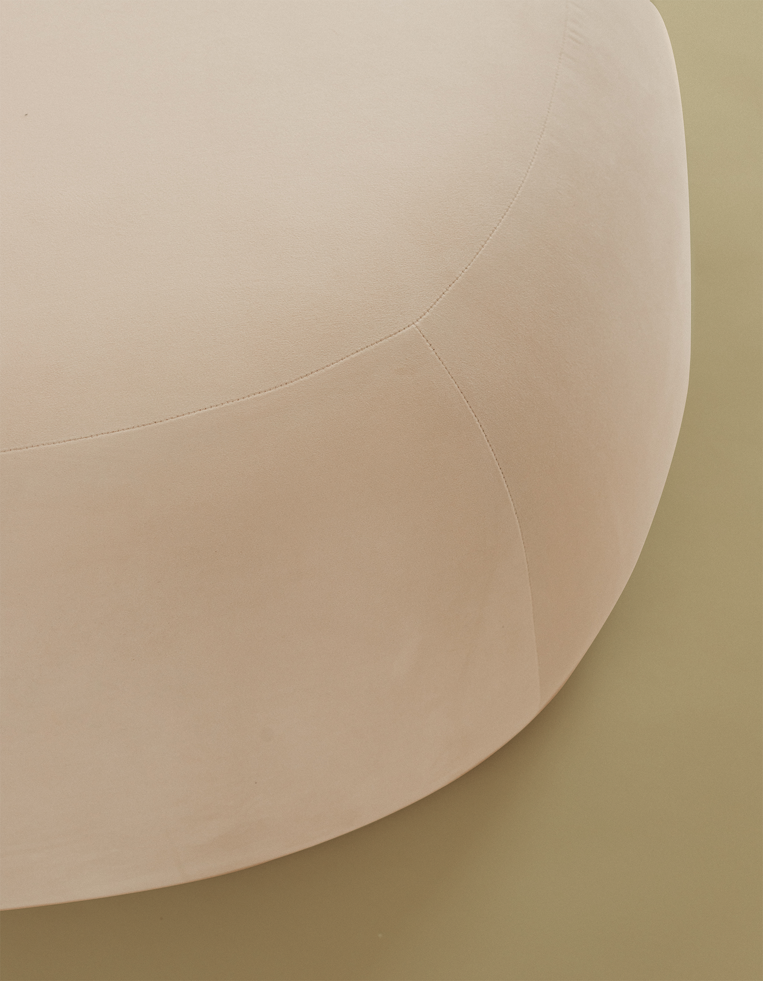 Close-up detail of an off-white pebble-shaped ottoman, showing the smooth upholstered surface and curved seam stitching on a beige studio background.