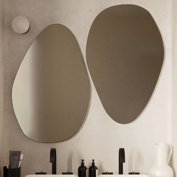 Two frameless pebble-shaped mirrors mounted above a double bathroom vanity with white vessel sinks and black faucets in a neutral-toned bathroom.