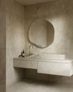Organic-shaped wall mirror with a slim stainless steel frame mounted above a stone vanity in a minimalist bathroom.