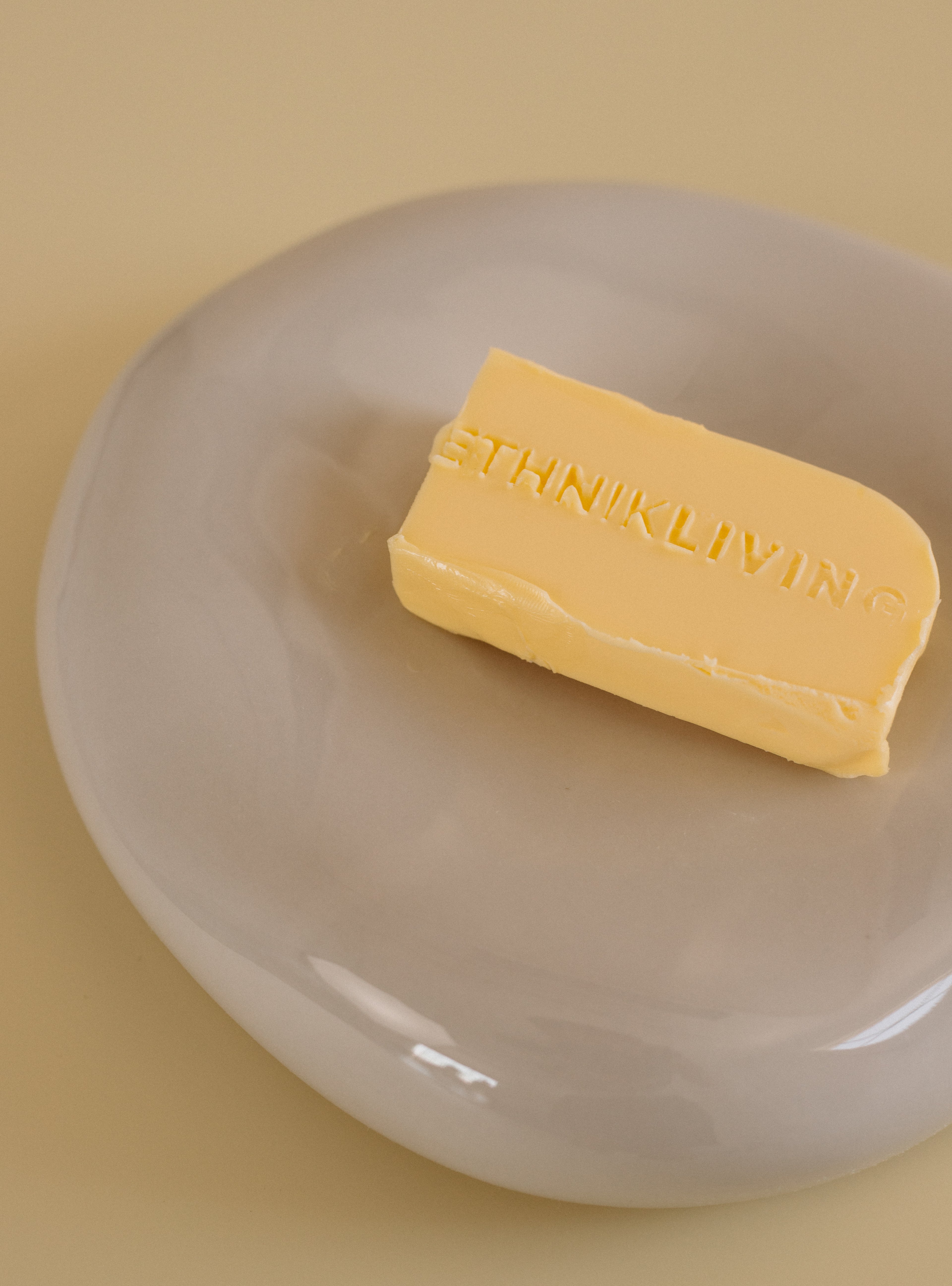 A rectangular stick of butter embossed with the text “ETHNIK LIVING,” placed on a round translucent dish resting on a light yellow side table.