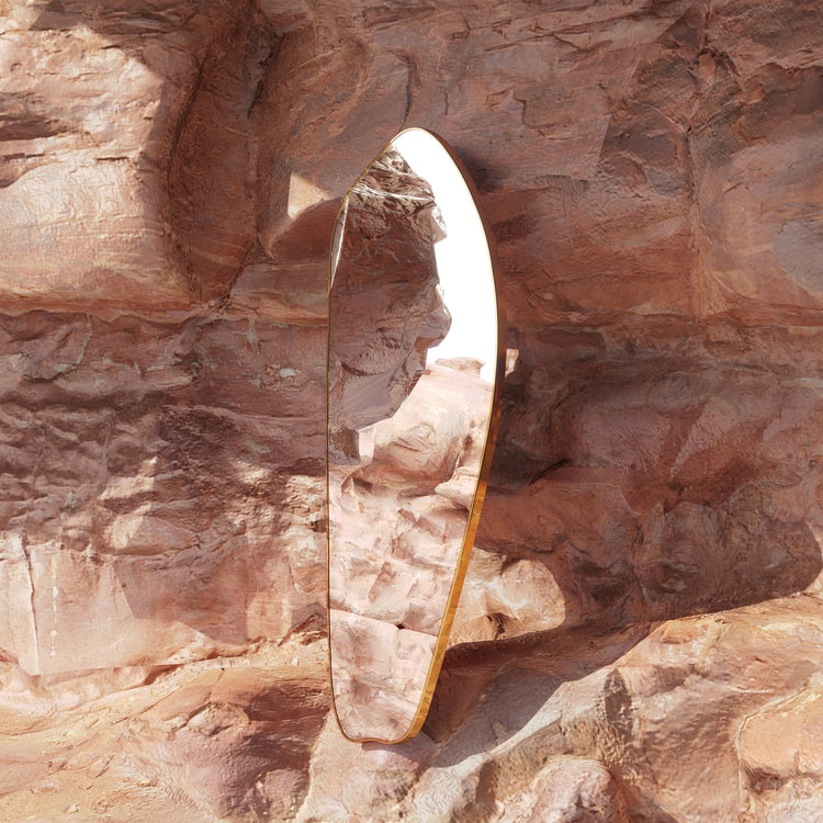 Asymmetrical brass-framed full length mirror placed against textured natural rock, reflecting the surrounding stone landscape.