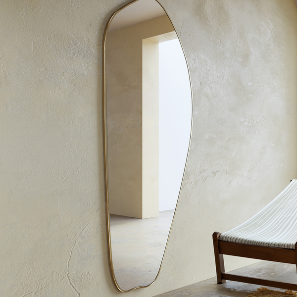 Asymmetrical brass-framed full length mirror mounted on a textured wall in a minimalist interior.