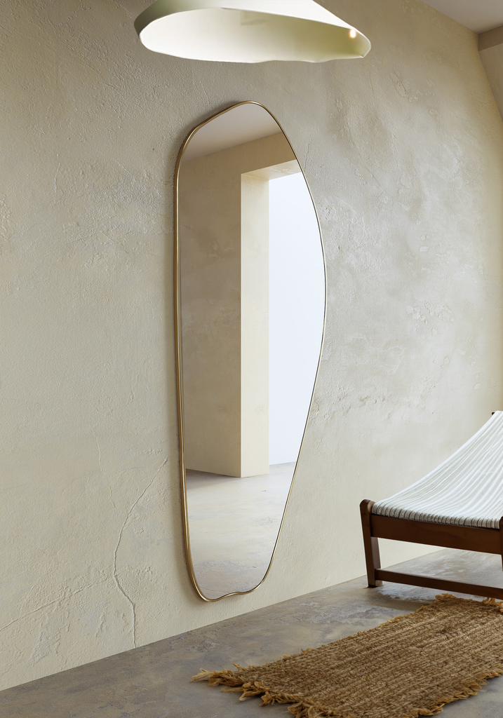 Asymmetrical brass-framed full length mirror mounted on a textured wall in a minimalist interior.