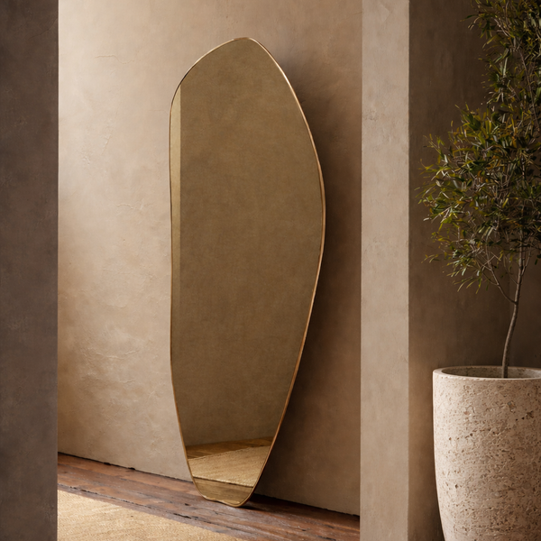 Organic-shaped floor mirror leaning against a textured wall, next to a potted plant in a minimalist interior.