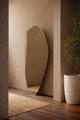Organic-shaped floor mirror leaning against a textured wall, next to a potted plant in a minimalist interior.
