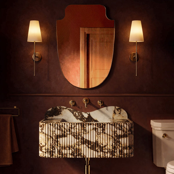 Moody bathroom with a sculpted wall mirror, brass sconces, and a marble pedestal sink against deep red walls.