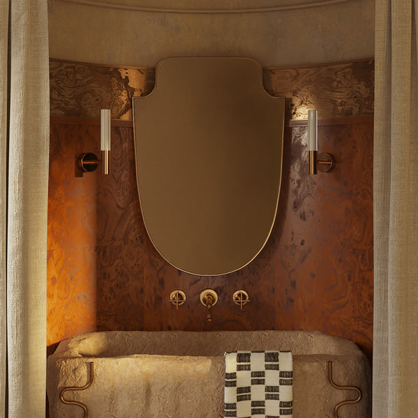 Shield-shaped wall mirror with a gold frame, mounted vertically above a bathroom vanity between two wall sconces.