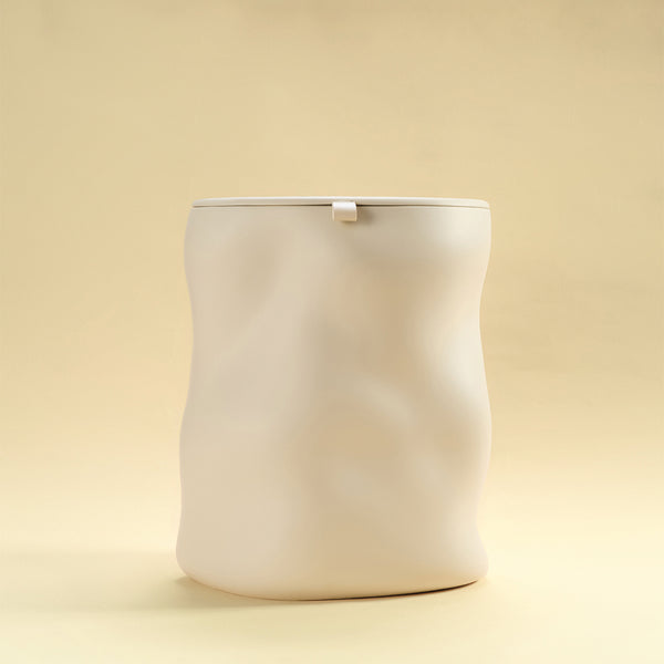 A beige sculptural dustbin with a fitted lid, photographed against a light yellow background.