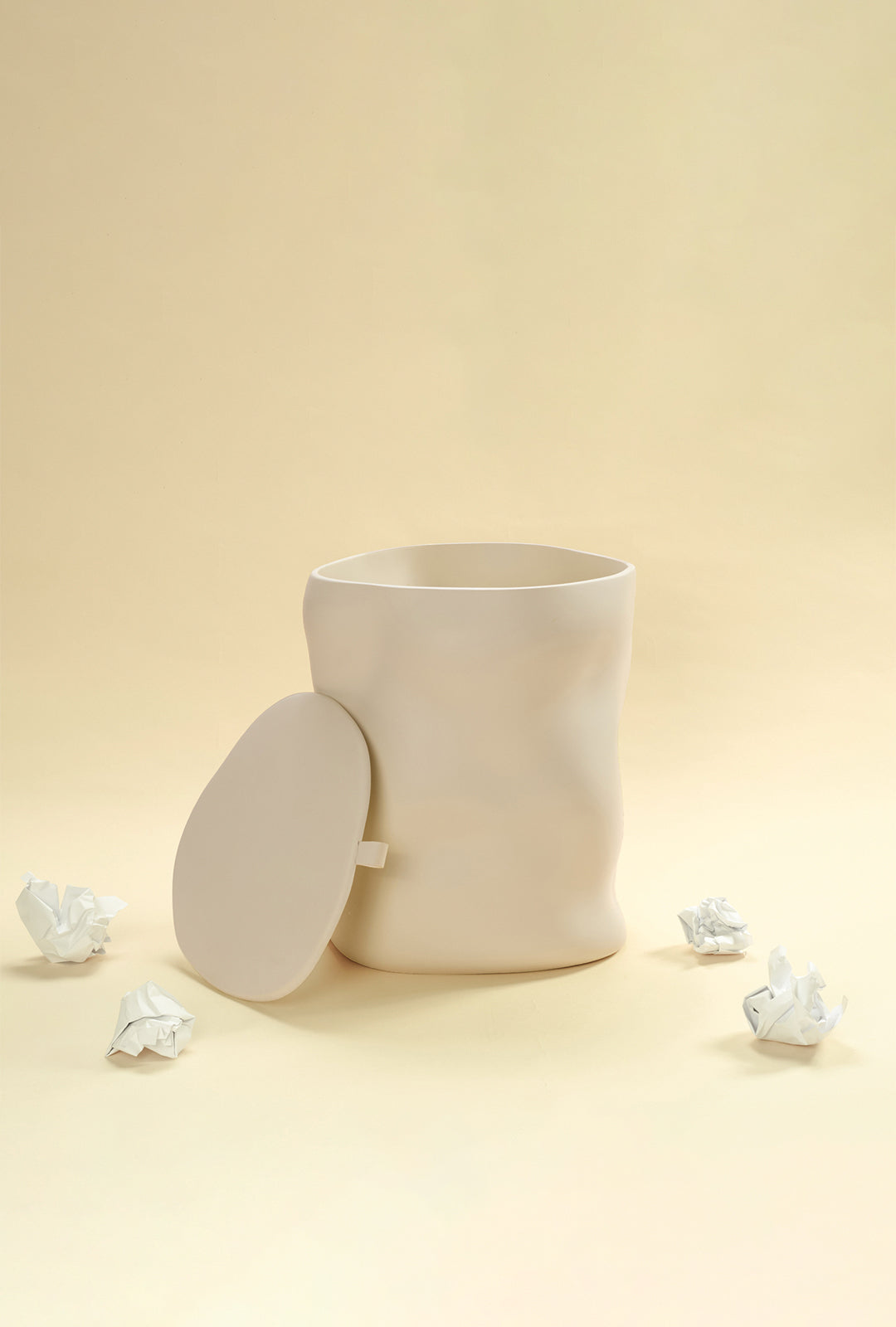 A beige sculptural dustbin shown with its lid removed and placed to the side, with several crumpled pieces of white paper on the floor around it, against a light yellow background.