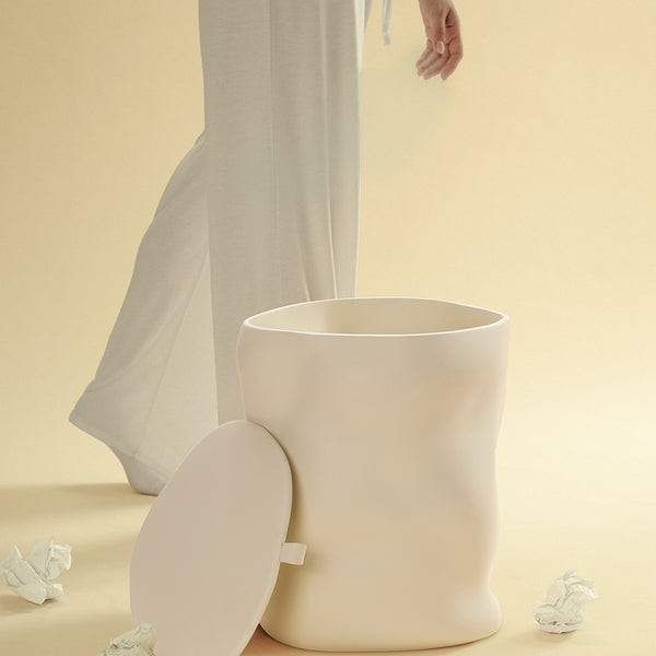 A beige sculptural dustbin shown with its lid placed to the side, as a person drops a crumpled piece of white paper into the open bin, against a light yellow background.
