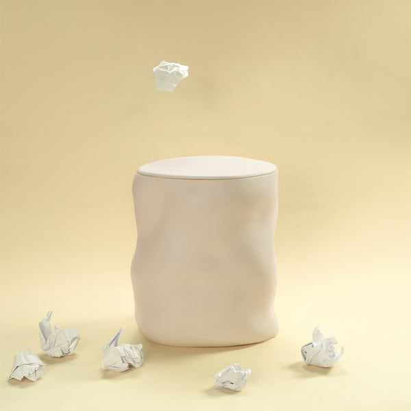 A beige sculptural dustbin placed against a light yellow background, with several crumpled pieces of white paper on the floor around it and one piece suspended above the bin.