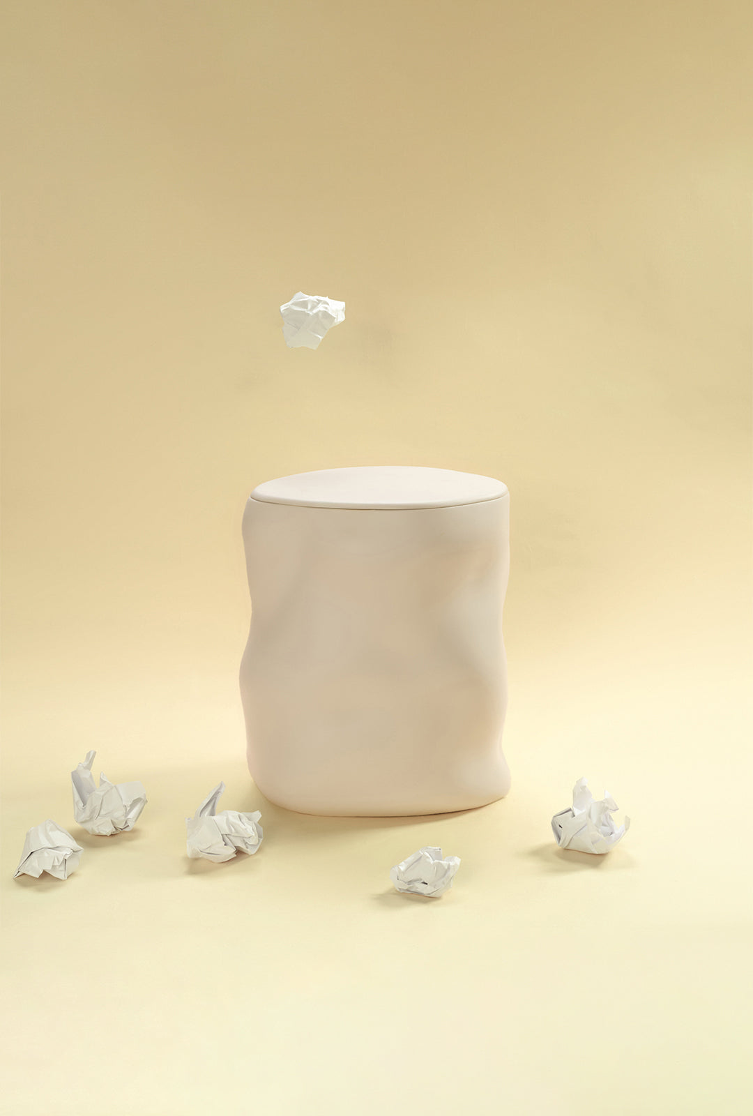 A beige sculptural dustbin placed against a light yellow background, with several crumpled pieces of white paper on the floor around it and one piece suspended above the bin.