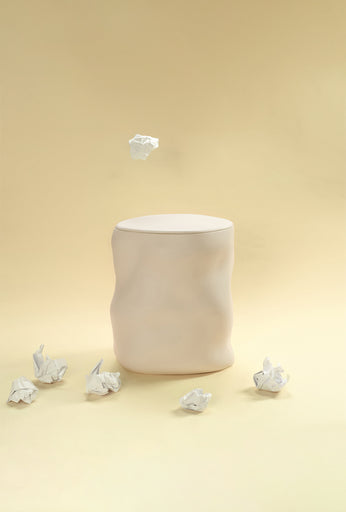 A beige sculptural dustbin placed against a light yellow background, with several crumpled pieces of white paper on the floor around it and one piece suspended above the bin.