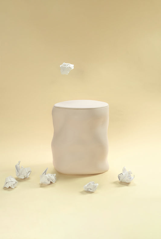 A beige sculptural dustbin placed against a light yellow background, with several crumpled pieces of white paper on the floor around it and one piece suspended above the bin.