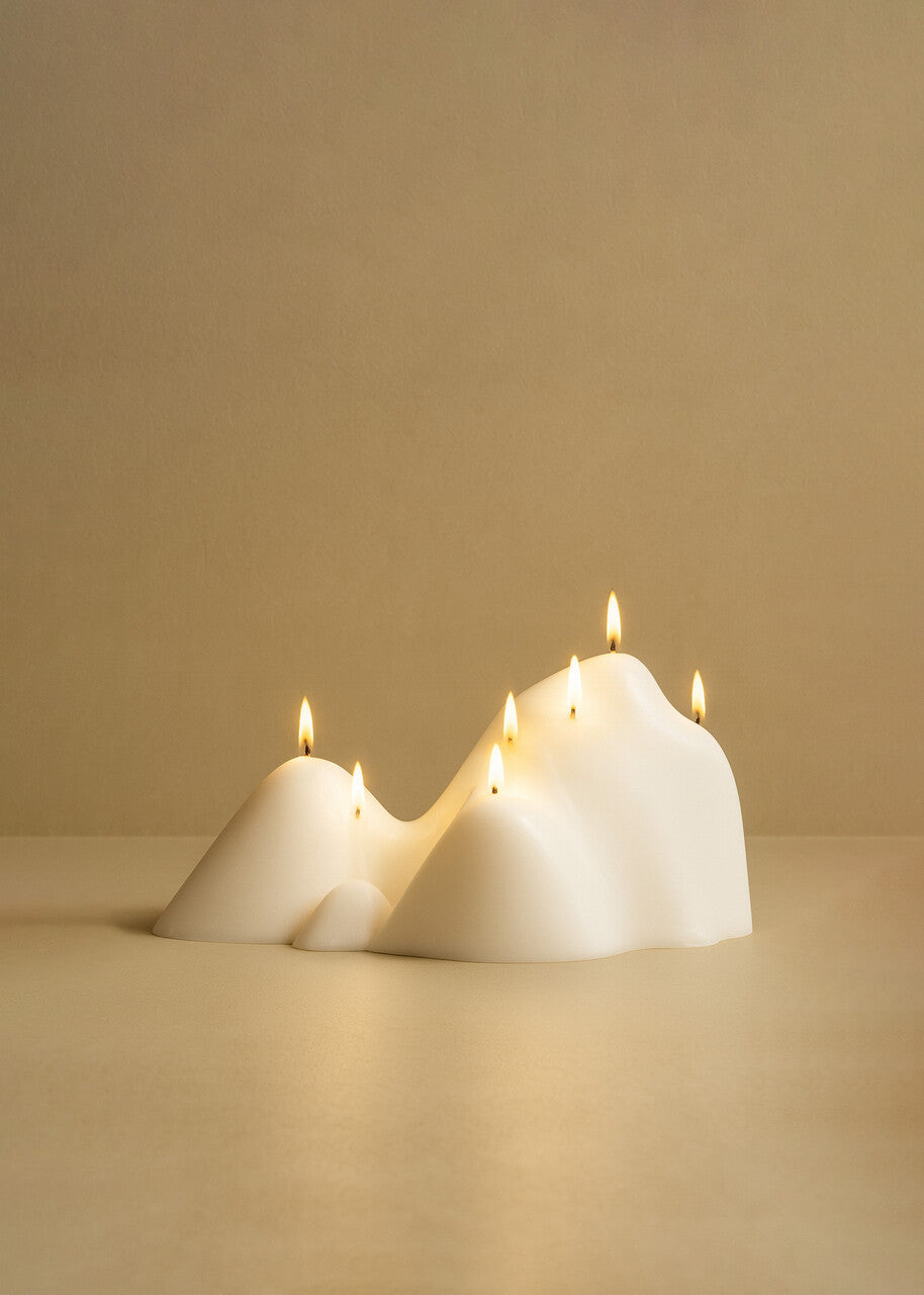 White sculptural candle with an organic, mountain-like form and multiple lit wicks, placed on a smooth surface against a warm beige background.