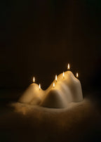 A sculptural white candle shaped like layered mountain peaks, with multiple small flames glowing softly against a dark background, creating a warm and atmospheric scene.